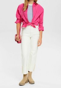 Kobieta w związanym różowym topie na paski, białych spodniach z wysokim stanem i szerokimi mankietami, beżowych botkach oraz niosąca różową torbę na ramię.