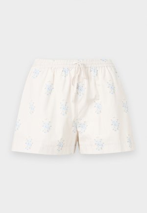 Femme debout portant un haut bleu à manches courtes, un short blanc à motifs floraux et des mules blanches à talons, sur un fond clair uni.