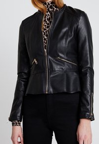 Giacca di pelle nera con un design corto, dotata di tasche con zip e accessori in argento, abbinata a un dolcevita con stampa leopardata sotto.