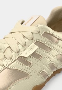 Κοντινή λήψη μπεζ δερμάτινου αθλητικού παπουτσιού με κρέμ κορδόνια, υφασμένη ραφή και λογότυπο της μάρκας στη γλώσσα και στο πλαϊνό πάνελ.