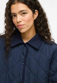 Mornarska podložena jakna s žametnim ovratnikom, ki ima zaponko spredaj in rombaste šive. Mehka tekstura in sproščen oblikovni slog.