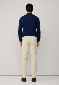 Suéter azul marino con parches negros en los codos, pantalones beige y zapatos marrón oscuro, visto desde atrás contra un fondo neutro.