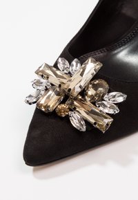 Chaussure en daim noire à bout pointu avec un regroupement de gros strass rectangulaires et ovales beiges et transparents à l'avant.