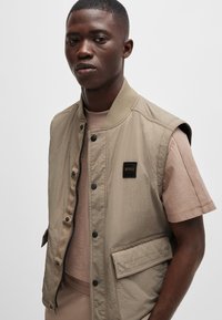 Beige ärmellose Jacke mit einem Reißverschluss vorne, Druckknöpfen und einem Brustlogo. Getragen über einem hellbraunen Kurzarmhemd. Glatte Textur.