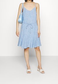 Ljusblå klänning med blommönster, justerbara axelremmar, V-ringning, knappdetaljer, dragsko i midjan och utsvängd kjol. Matchad med vita klackskor med blockklack och en blå axelväska.