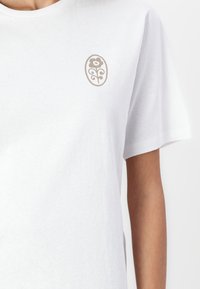 T-shirt en coton blanc à manches courtes, avec un emblème floral beige en forme d'oval sur la poitrine. Texture douce, coupe décontractée.