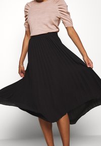 Zwarte geplooide rok met hoge taille, gemaakt van zacht materiaal, gecombineerd met een lichtbruine top met korte mouwen en geplooide details.