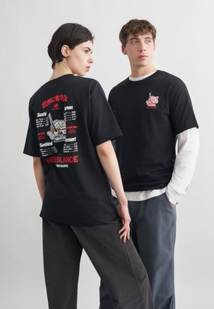 Δύο μοντέλα φορούν μαύρα t-shirts της New Balance με θέμα το σούσι· το ένα κοιτάζει προς τα πίσω και δείχνει το σχέδιο με το μενού, ενώ το άλλο κοιτάζει πλάγια φορώντας ένα μακρυμάνικο με στρώσεις.