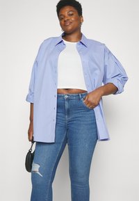 Lys blå oversized skjorte med knapper og oppbrettede ermer, brukt over en hvit cropped topp, sammen med blå smale jeans med et hull.