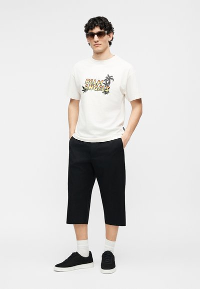 Άνδρας που φοράει γυαλιά ηλίου, λευκό t-shirt "Palm Angels" με γραφικό φοίνικα, μαύρο παντελόνι φαρδύ και κοντό, λευκές κάλτσες και μαύρα αθλητικά παπούτσια.
