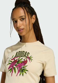 Beżowa bawełniana koszulka z kolorową grafiką różowych papug i zielonych liści, z napisem "ADIDAS" w pogrubionych czarnych literach powyżej.