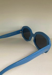 Lunettes de soleil octogonales bleues avec des montures en plastique épais, des verres foncés et une texture lisse. Présente un marquage de la marque sur la branche.