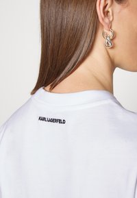 Valkoinen puuvillapaita, jossa on pyöreä pääntie, takana musta "KARL LAGERFELD" kirjailu. Mallilla on päällä hopeiset ja kultaiset korvakorut.