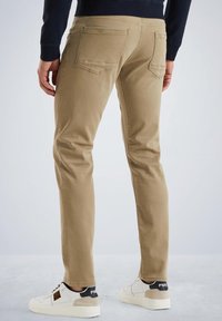 Pantaloni slim-fit color khaki con una texture liscia, due tasche posteriori e un dettaglio di cucitura, abbinati a sneakers bianche con accenti neri.