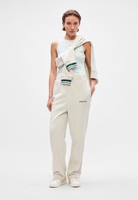 Ανοιχτό μπλε αμάνικο μπλουζάκι με λογότυπο, σε συνδυασμό με κρεμ παντελόνια φόρμας που διαθέτουν ζώνη. Ελαφρύ πουλόβερ ριγμένο στους ώμους με πράσινες λεπτομέρειες.