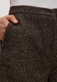 Hand in de voorzak van bruine en grijze geruite textuurbroek met een knoopsluiting in de taille.