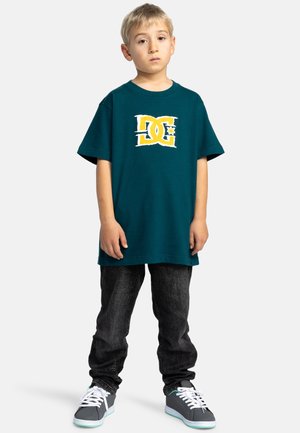 Türkises Baumwoll-T-Shirt mit einem weißen und gelben Logo, kombiniert mit dunklen Jeans und grauen Sneakers mit weißen Schnürsenkeln und türkisfarbenen Akzenten.