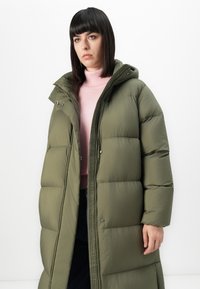 Kvinde med lige sort hår iført en lang olivengrønn pufferjakke over en lyserød turtleneck sweater mod en hvid baggrund.