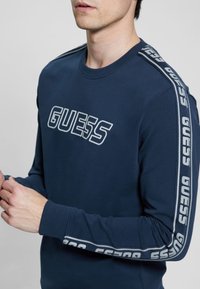 Felpa blu marina con il logo "GUESS" sul davanti. Le maniche lunghe presentano strisce a motivo lungo i lati con dettagli bianchi. Tessuto in misto cotone.
