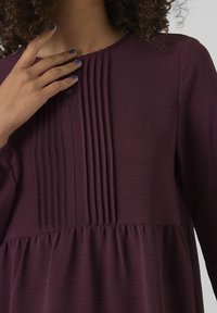 Bordeauxfarbenes Oberteil mit vertikalen Falten am Ausschnitt, strukturiertem Stoff und ausgestellter Silhouette. Lange Ärmel und runder Kragen.
