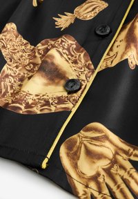 Cămașă neagră cu imprimeuri grafice aurii reprezentând mâini și ananasuri, cu nasturi negri și un accent cu fermoar auriu de-a lungul marginii.