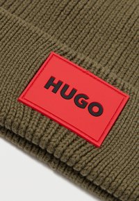 Beret tricotat de culoare verde măslin, cu textură în dungi, având un patch rectangular roșu pe care este afișat în mod proeminent logo-ul negru "HUGO".