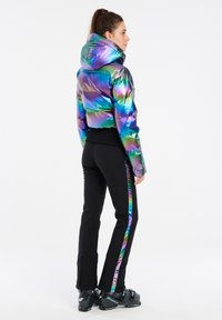 Iriserende opblaasjasje met een capuchon, met een meerkleurig patroon. Combinatie met zwarte broeken met een reflecterende, kleurrijke streep.