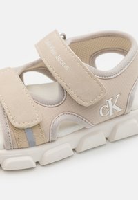 Beige sportsandal med borrelås, teksturert overdel, polstret såle og "CK"-logo på siden; har grå akcentstripe på overdelen.