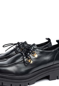 Zapatos de cuero negro con una textura suave, que presentan ojales dorados, cordones negros y una suela negra gruesa con un patrón de agarre.