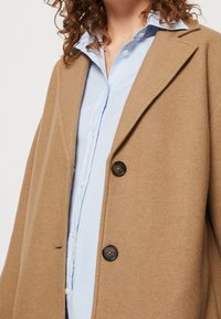 Kameluld-blanding frakke med hakket revers, udstyret med to sorte knapper og en blød, tekstureret finish. Båret over en lyseblå skjorte med knapper.