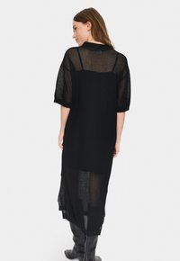 Rochie tricotată neagră cu mâneci scurte, textură de tricot deschis, lungime mare și tăietură pe margine. Prezintă o croială lejeră și detalii cu dungi.