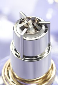 Silver parfymflasklock med ett facetterat, gemliknande topp och vinklade detaljer. Basen har en ring i guldfärg. Slät metallisk yta.