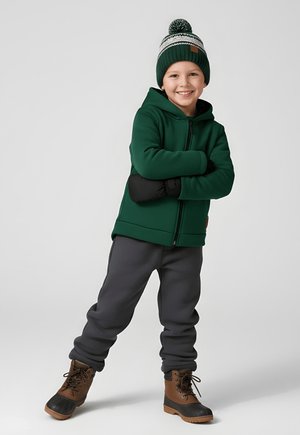 Uśmiechnięte dziecko w zielonej bluzie z kapturem, dzianej czapce z pomponem, czarnych rękawiczkach, szarych spodniach dresowych i brązowych zimowych butach, stojące z założonymi ramionami.