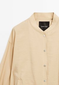 Ljusbeige lättviktsjacka med mandarinliknande krage, tryckknappsstängning och texturerad tyg. Inuti finns en etikett som visar "Massimo Dutti."