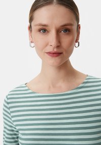 Gestreiftes Langarmshirt in Hellgrün und Blau mit einem runden Ausschnitt. Das Modell trägt silberne Creolen und hat eine glatte Hautstruktur.