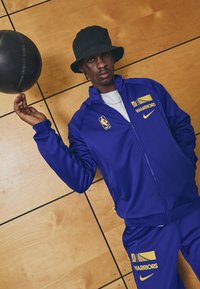 Karališkai mėlynas treniruočių kostiumas su geltonais akcentais, su "WARRIORS" logotipu, NBA emblemą ir juodu kibiru, laikantis juodą krepšinį.