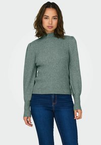 Ribbad långärmad polotröja i dämpad grön, med figurnära passform och subtila puffade axeldetaljer, parat med mörkblå jeans.