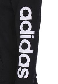 Sweat-shirt noir avec le texte "adidas" en blanc imprimé verticalement sur la manche. Le matériau semble doux avec une texture lisse.