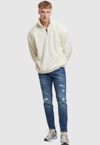Pull en polaire blanc avec un col haut et une fermeture éclair à l'avant, associé à un jean bleu déchiré et des baskets argentées.