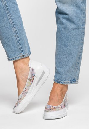 Füße, die weiße Slip-On-Schuhe mit floralen Mesh-Details tragen, kombiniert mit hellblauen, hochgekrempelten Jeans vor einem schlichten Hintergrund.