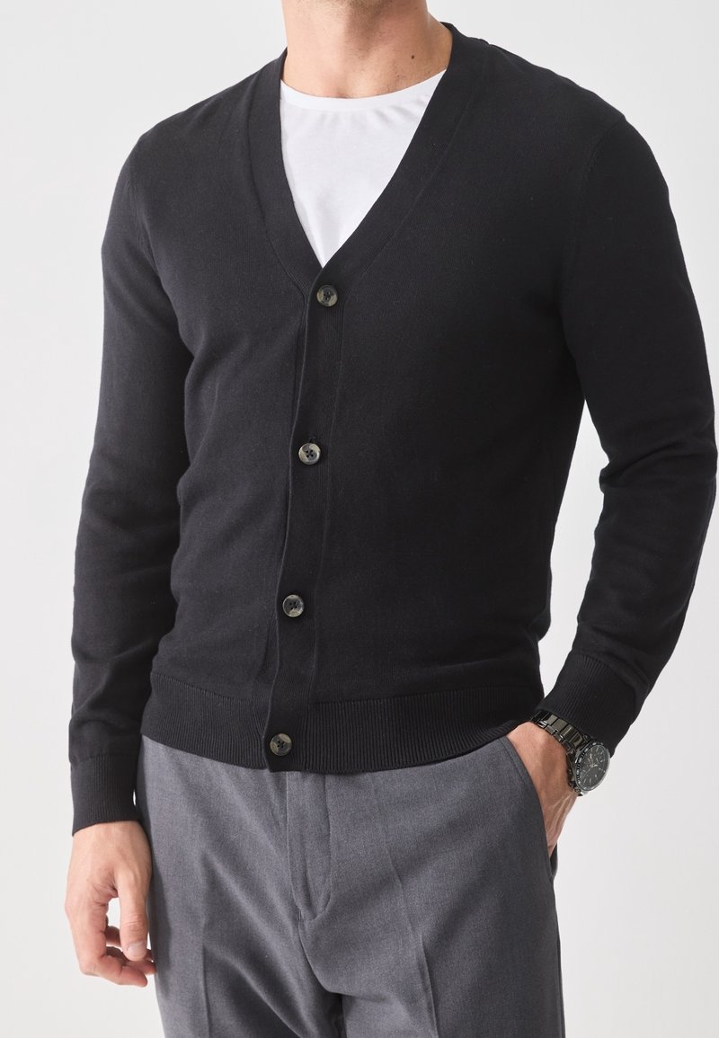 Homme portant un cardigan noir boutonné sur une chemise blanche avec un pantalon gris et une montre-bracelet, main dans la poche, sur un fond uni.