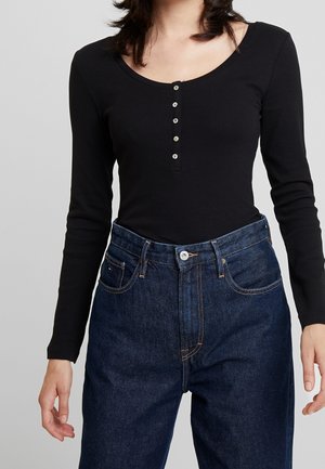 Femme portant un haut noir à manches longues côtelé avec des boutons et un jean en denim bleu foncé taille haute sur un fond gris uni.