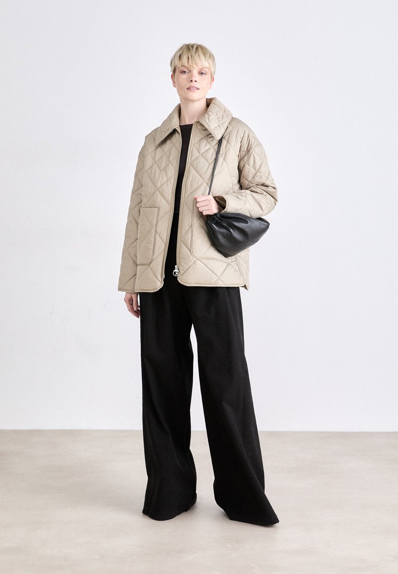 Beige gesteppte Jacke mit großem Kragen, Fronttaschen und Reißverschluss, kombiniert mit weiten schwarzen Hosen und einer kleinen schwarzen Tasche.