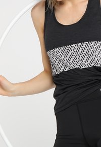 Černý tílko s geometrickým bílým vzorem přes hruď. Vyrobeno z hladkého materiálu, s volným střihem a zakřiveným lemem.