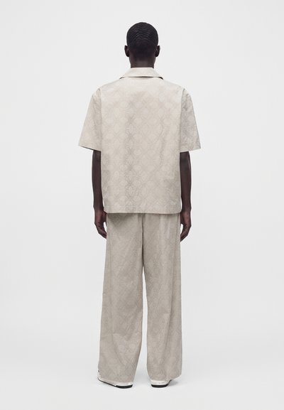 Personne aux cheveux courts et foncés portant une chemise beige à manches courtes et un pantalon ample et large assorti, debout de dos sur un fond uni.
