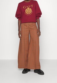 Sudadera burdeos con logo dorado, combinada con pantalones de cuadros naranjas y marrones de pierna ancha. Unos elegantes zapatos de plataforma negros completan el look.