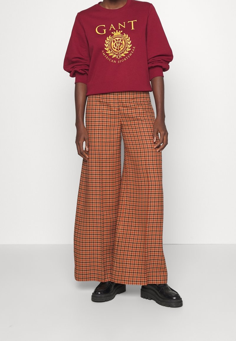 Sudadera burdeos con logo dorado, combinada con pantalones de cuadros naranjas y marrones de pierna ancha. Unos elegantes zapatos de plataforma negros completan el look.