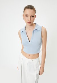 Top cropped sin mangas de canalé azul claro con cuello, combinado con pantalones blancos de cintura alta. Textura suave, diseño ajustado.