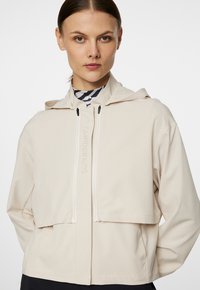 Beige cropped hoodie met een gladde textuur, twee voorzakken en witte ritsdetails. Heeft een reliëflogo aan de voorkant.