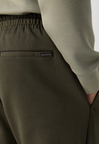 Olivgröna sweatpants med mjuk textur, elastisk midja och en bakficka med en liten svart varumärkesetikett.